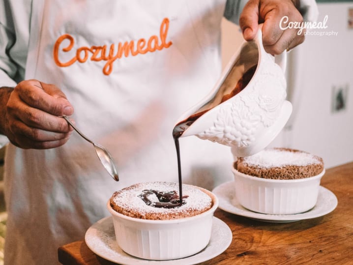 Chef in a Cozymeal apron pouring rich chocolate sauce over individual soufflÃ©s in white ramekins.