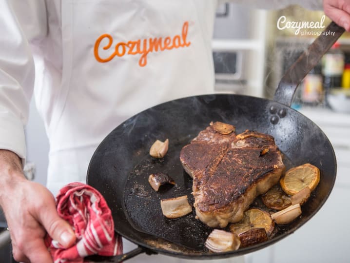 Chef in Cozymeal apron holding cooked steak in a pan