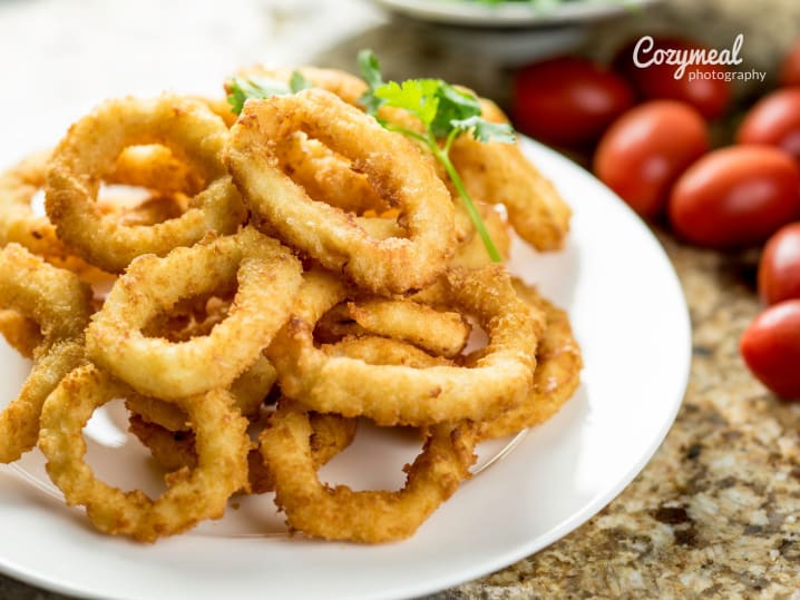 A white plate filled with crispy golden fried calamari rings, served with lemon wedges and cherry tomatoes