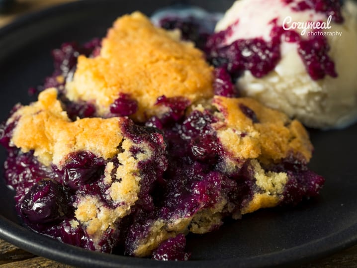 Blueberry cornbread cobbler with vanilla ice cream â€“ A scoop of warm blueberry cornbread cobbler served with a scoop of vanilla ice cream on a black plate.