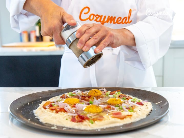 chef in Cozymeal apron adding toppings to carbonara-style pizza