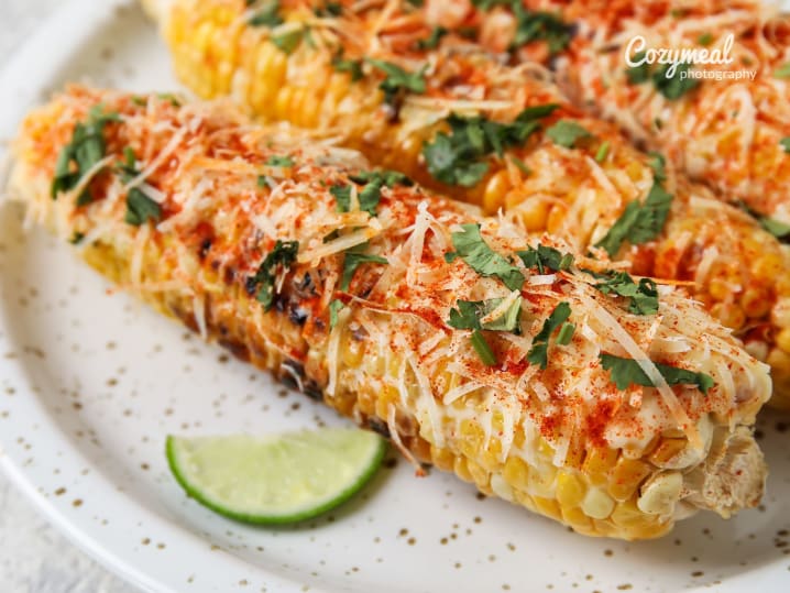 Grilled elote corn garnished with cheese, herbs, and lime wedges