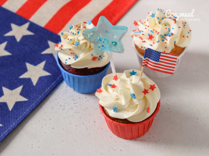 Fourth of July cupcakes with patriotic toppers â€“ Three vanilla cupcakes with white frosting and star-shaped sprinkles, one topped with an American flag and another with a blue candy star.