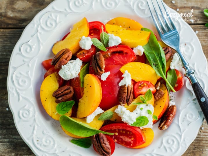 Peach and feta salad with greens and pecans â€“ A summer salad with sliced peaches, feta, leafy greens, roasted red peppers, and pecans on a white plate.
