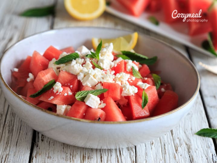 Watermelon salad with feta and mint â€“ A bowl of cubed watermelon tossed with crumbled feta and fresh mint, served with a lemon wedge on a rustic white table.