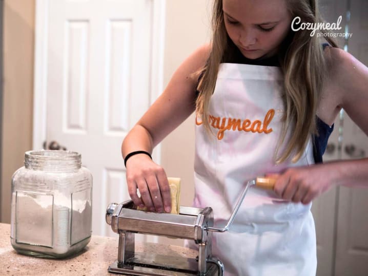 a young teenage girl wearing Cozymeal apron is making fresh pasta using pasta machine