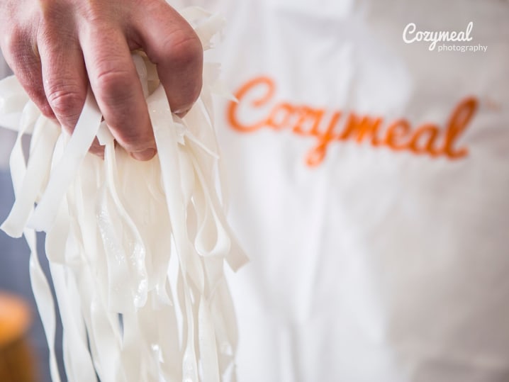 Chef holding rice noodles â€“ A close-up of a chef in a â€œCozymealâ€ apron holding a bundle of uncooked white rice noodles.
