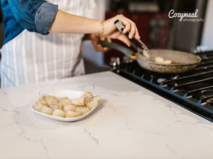 Chef cooking scallops on stovetop â€“ A person searing scallops in a pan on the stove, with a plate of raw scallops on the marble counter nearby, mid-prep during the Bastille Day Feast cooking class.