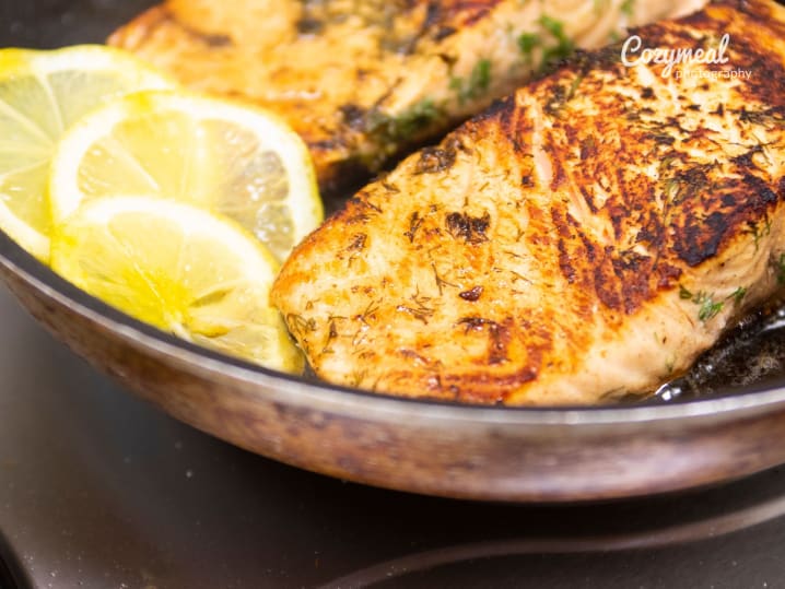 Cooking salmon fillets in skillet â€“ Salmon fillets searing in a skillet with lemon slices, seasoned with herbs and spices.