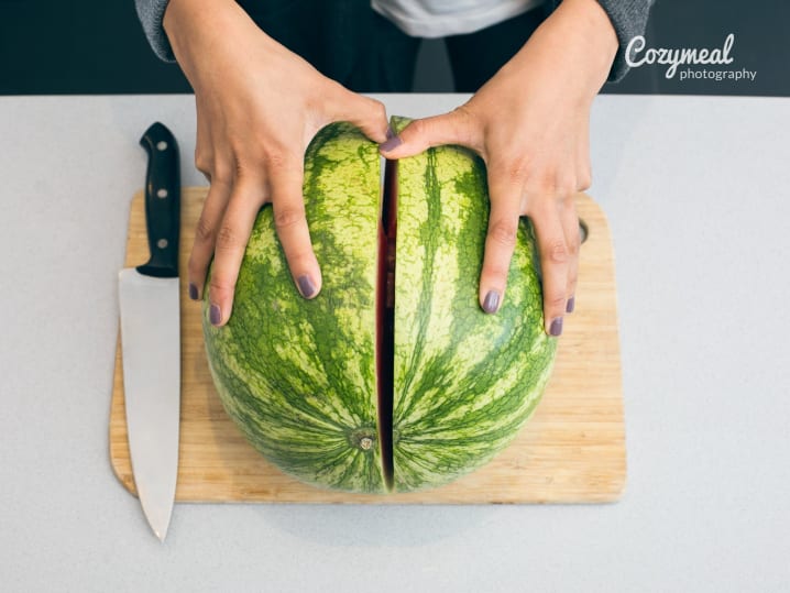 Cutting a whole watermelon in half â€“ Overhead view of hands slicing a whole watermelon in half on a wooden cutting board with a chefâ€™s knife beside it.