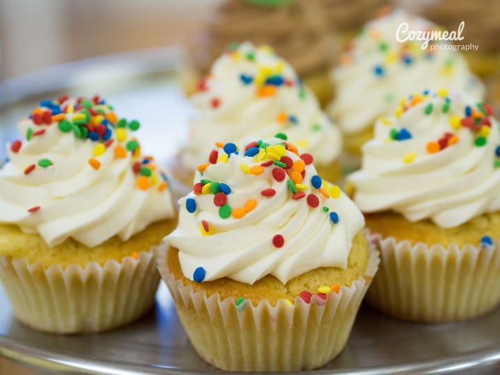 Close-up of vanilla cupcakes with white frosting and colorful sprinkles, arranged on a metal tray.