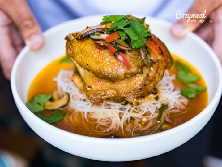 Grilled chicken breast over chili broth and noodles â€“ A bowl of grilled chicken served atop rice noodles in a rich chili-infused broth, garnished with fresh herbs.