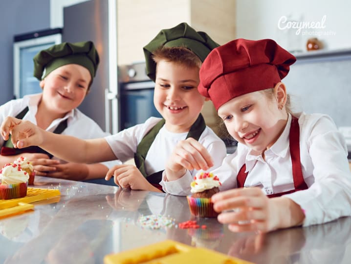 Group of kids wearing colorful chef hats, decorating cookies with icing and sprinkles at a table.