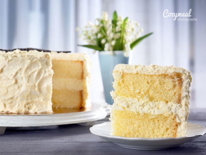 Slice of layered vanilla cake with white frosting, served on a white plate with a cake stand in the background.