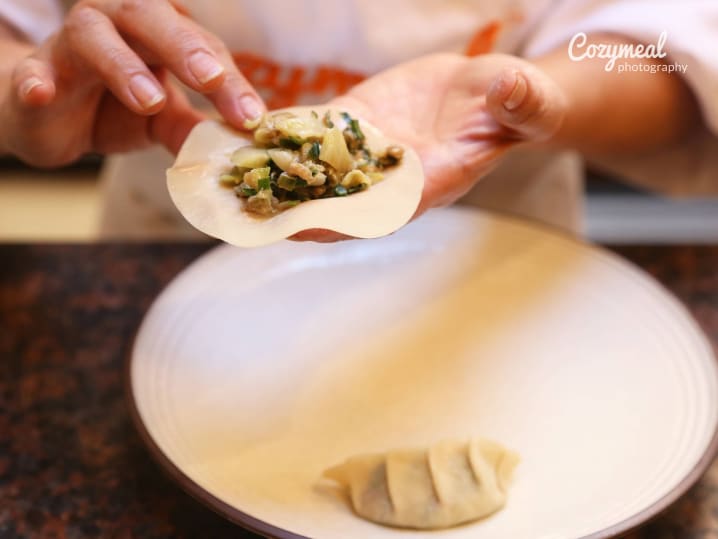 Folding dumplings with vegetable filling â€“ A close-up of hands shaping a dumpling with a green filling over a round white plate.