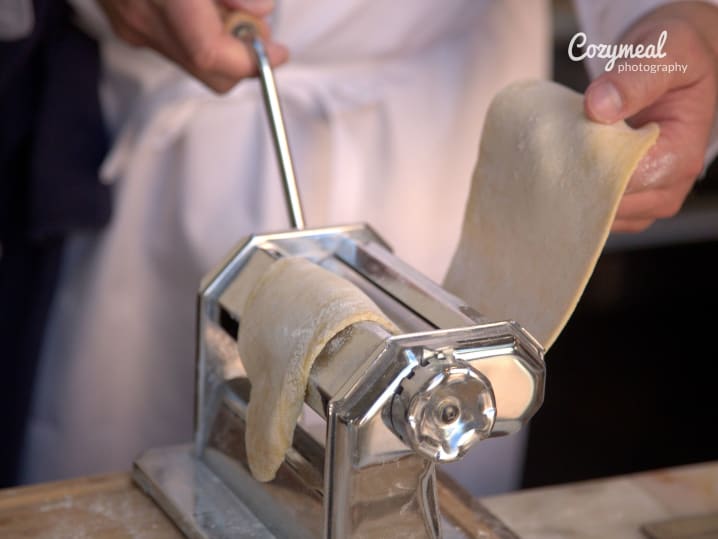 Rolling pasta dough through pasta machine â€“ Close-up of a person feeding sheets of pasta dough through a hand-cranked pasta roller.