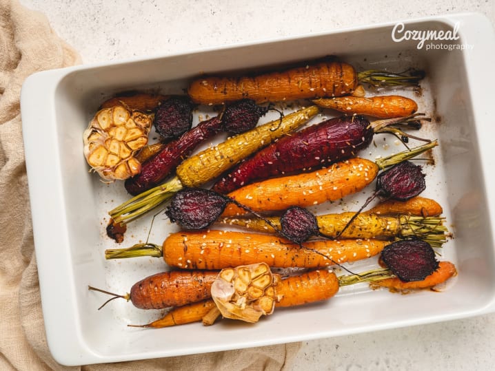 Rainbow carrots roasting in a white pan â€“ Multicolored carrots roasting in a white baking dish, garnished with herbs and resting on a beige towel.