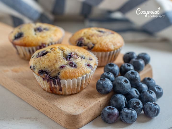 three blueberry muffins on a wooden board next to a batch of fresh blueberries