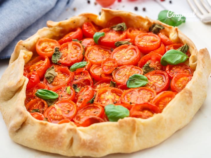 Rustic tomato tart with basil â€“ A golden galette-style tart filled with cherry tomatoes and topped with fresh basil leaves on a white surface.