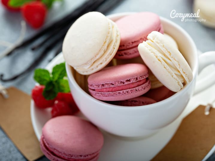 Vanilla and raspberry macarons in teacup â€“ A mix of white and pink macarons stacked in a teacup with fresh raspberries and mint on the side.