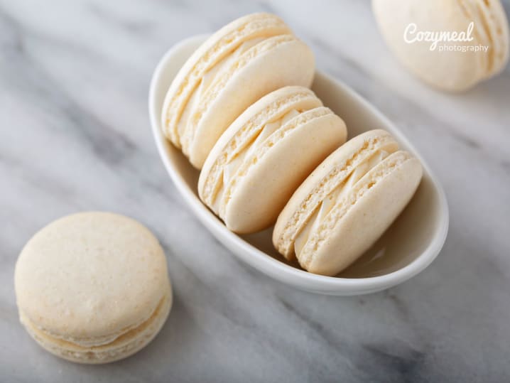 White macarons on marble surface â€“ Three vanilla macarons placed neatly on a white marble slab with a shallow dish of more in the background.
