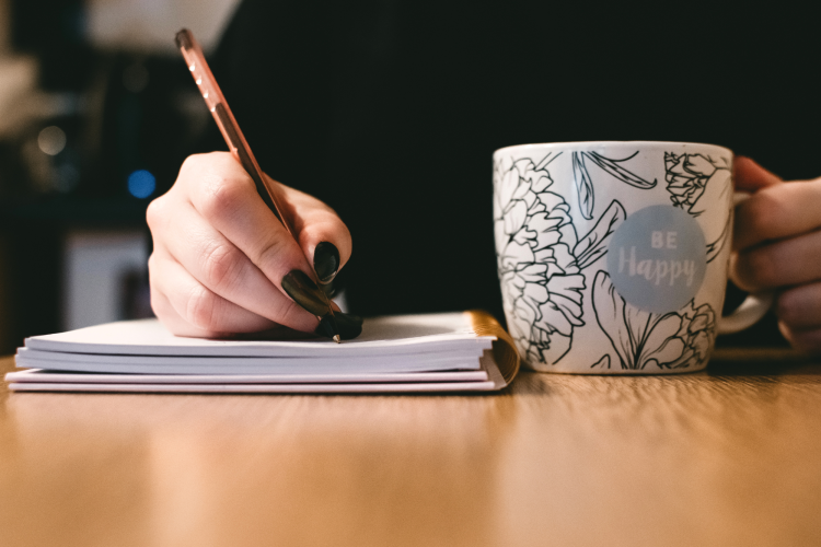 handwriting in notebook while holding painted mug that says