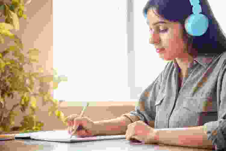 young woman writing notes while listening to audiobook on headphones