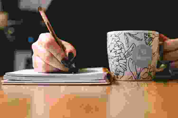 handwriting in notebook while holding painted mug that says 