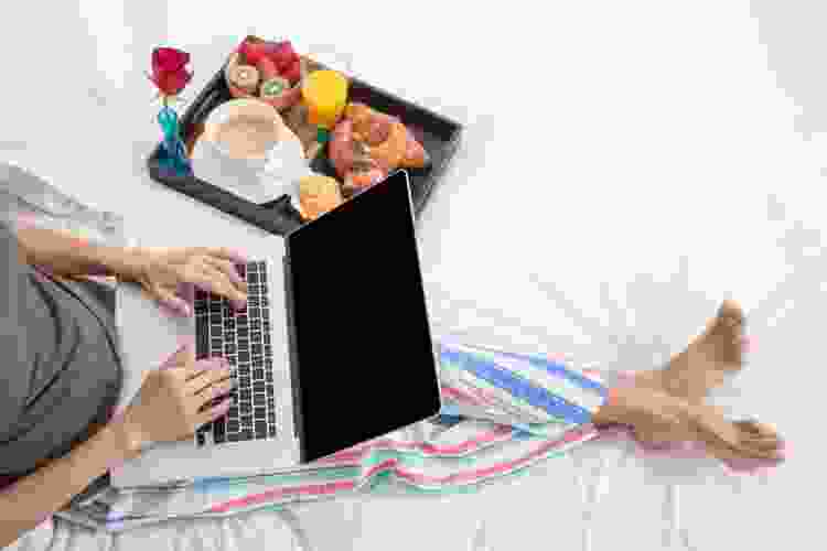 person laying in bed with tray of brunch foods and laptop in their lap