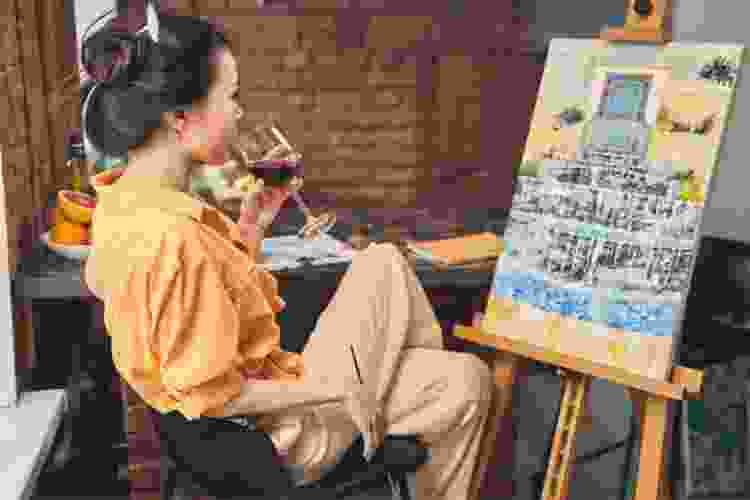 woman sipping wine while painting canvas