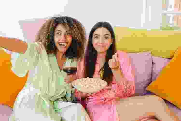 two friends eating popcorn and drinking wine on a couch