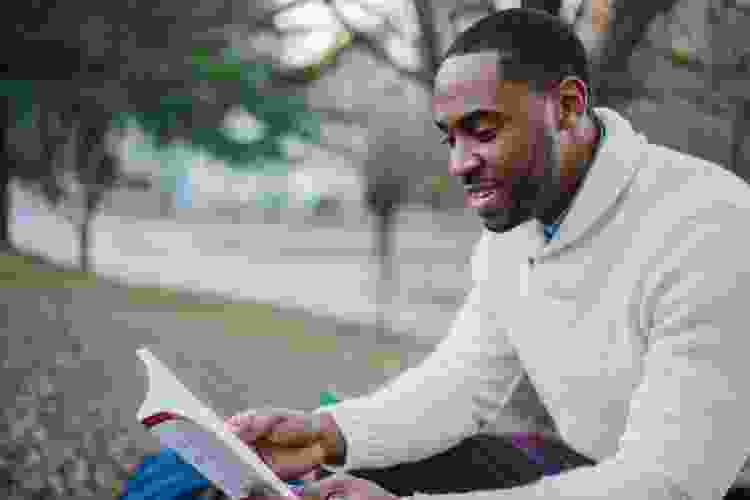 man reading book outside on a park bench