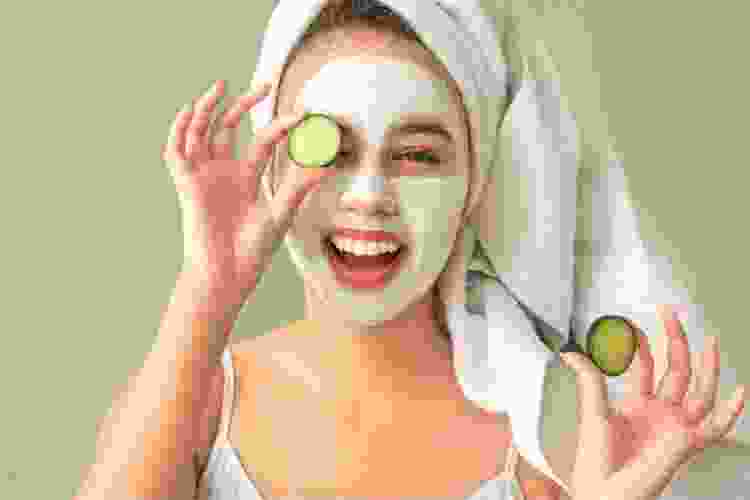 young woman in mirror wearing face mask and holding cucumber slice