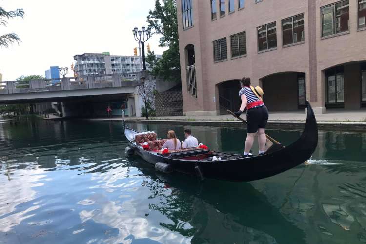 two people ride in a gondola while a female gondolier in striped clothes steers