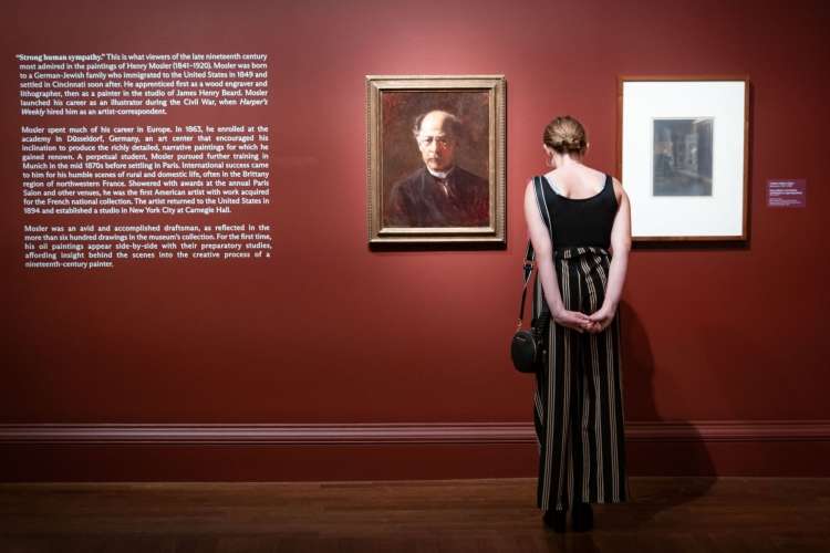 a woman stands with hands behind back, looking at a painted portrait in a museum