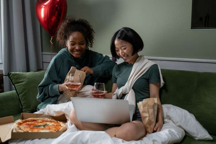 two women watch a laptop on the sofa while eating pizza and drinking beer