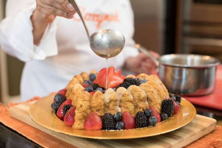 a chef in a Cozymeal apron drizzles white sauce over a fruit and ladyfingers cake