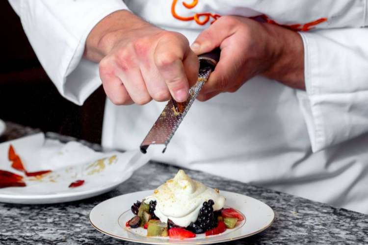 a chef in Cozymeal apron grates white chocolate over a fruity dessert