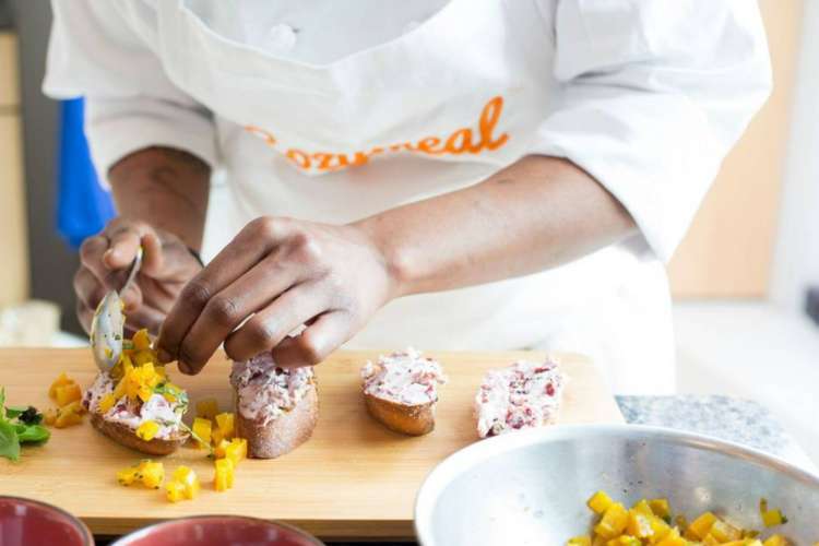 A chef wearing a Cozymeal apron spoons tuna salad onto bred slices.