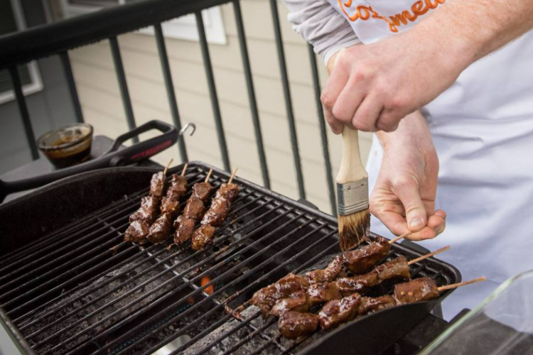 a chef wearing a Cozymeal apron brushes sauce on barbecue kebabs