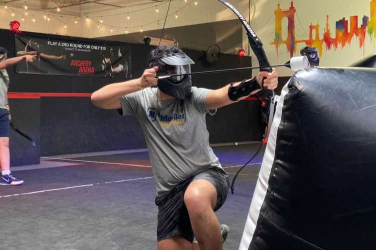 a helmeted archery player kneels down with a foam-tipped arrow pulled back 