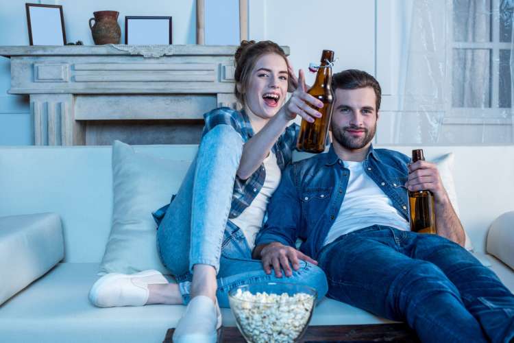 a man and woman watch TV on a sofa while eating popcorn