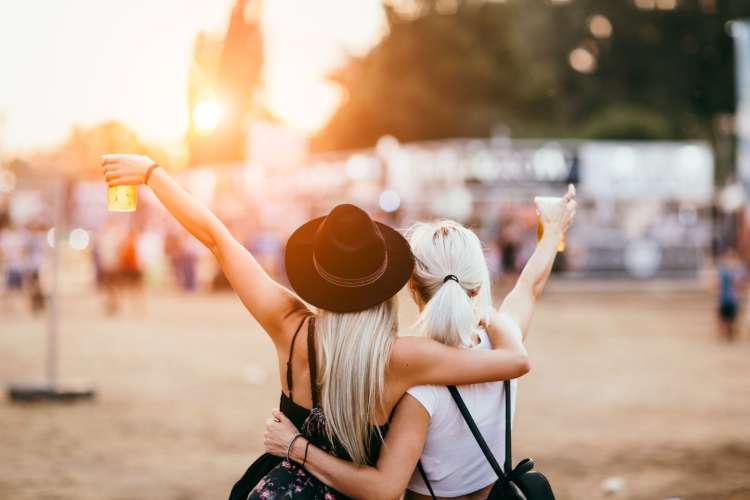 two women at a music festival hug and raise drinks 