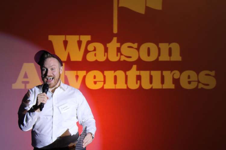 man speaking in front of a red background that reads Watson Adventures