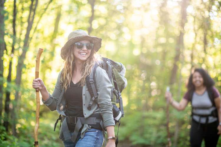 a smiling woman with sunglasses, hat and backpack while hiking