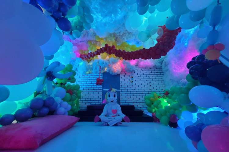 A person in a unicorn costume sitting in a white brick room full of balloons