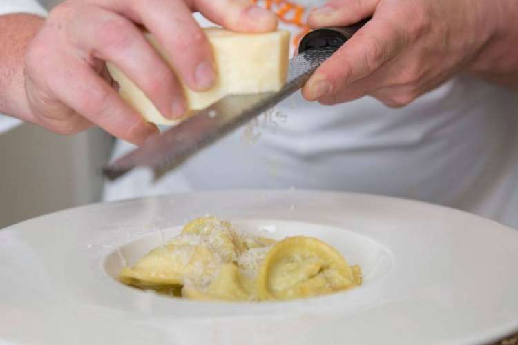 A chef in a Cozymeal apron grates cheese over a pasta dish.