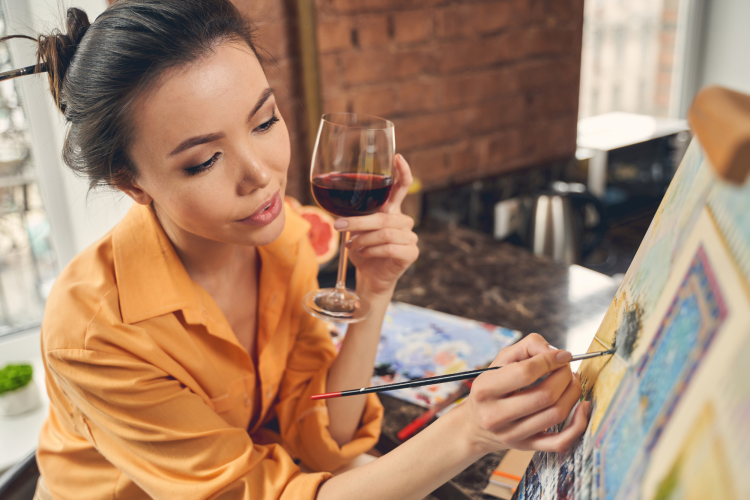 woman painting on a canvas while drinking red wine