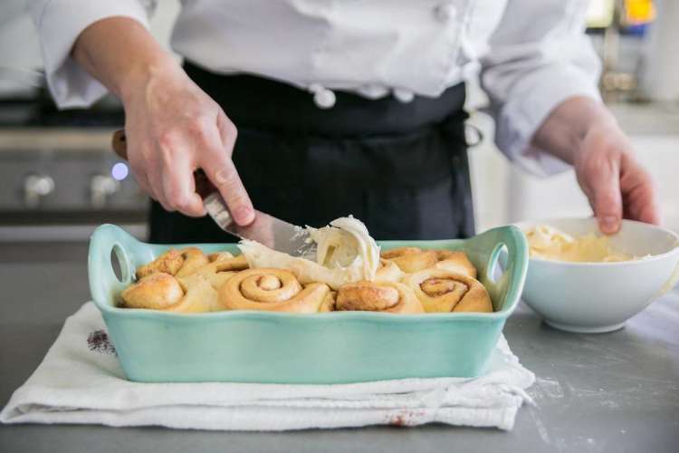 chef smearing cream cheese icing over homemade cinnamon rolls