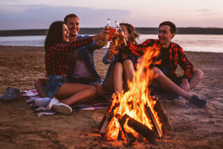 friends toasting beers at a beach bonfire at dusk